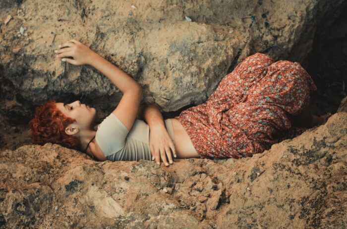 A young red haired feminine presenting person wearing a grey t-shirt top and a long multicolored floral skirt lying under the edge of a very large boulder pressing down on-top of them.